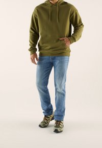 Felpa con cappuccio verde oliva, tasca frontale, vestibilità comoda; jeans azzurri chiari; scarpe con motivo camouflage con suole nere e lacci marroni.