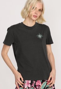 Czarna bawełniana koszulka typu t-shirt z krótkim rękawem, z małym haftowanym wzorem gwiazdy w kolorze turkusowym i białym po lewej stronie klatki piersiowej.