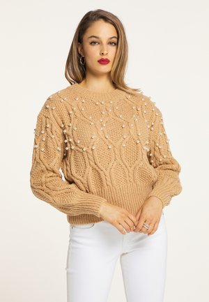 Donna con capelli fino alle spalle che indossa un maglione color sabbia decorato con perle e pantaloni bianchi, in piedi con le mani giunte davanti.