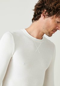 Camicia bianca a maniche lunghe con texture a coste e scollatura rotonda, caratterizzata da un sottile dettaglio di punto incrociato al colletto.
