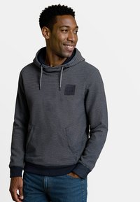 Man in een marineblauwe en grijze gestreepte hoodie met een voorkantzak en trekkoord, glimlachend en kijkend naar rechts.