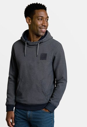 Man in een marineblauwe en grijze gestreepte hoodie met een voorkantzak en trekkoord, glimlachend en kijkend naar rechts.