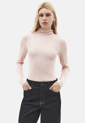 Kvinde med langt blondt hår iført en lyserød ribbet rullekrave og sorte højtaljede jeans med hænderne i lommerne.