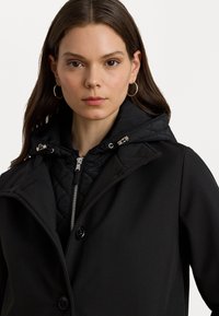 Manteau noir à l'extérieur avec une couche intérieure matelassée, doté d'une capuche, d'une fermeture éclair et de deux boutons. Texture lisse, coupe ajustée et design minimaliste.
