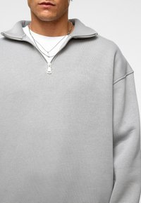 Lichtgrijze ritsvest met hoge kraag en lange mouwen, gemaakt van zachte stof. Voorzien van een voorkant rits en een relaxte pasvorm.