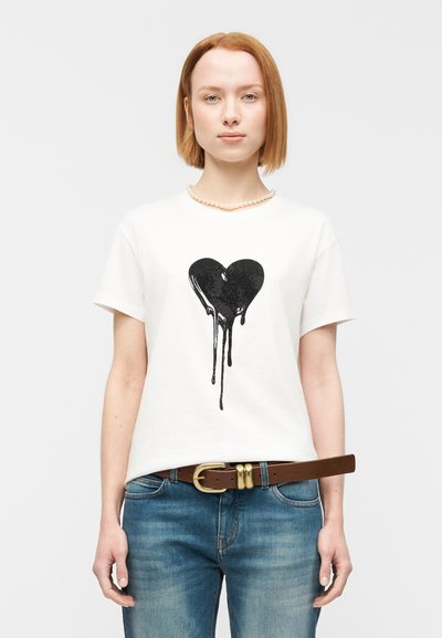 Άσπρο βαμβακερό t-shirt με μαύρο γραφικό καρδιάς που στάζει στο κέντρο. Το μοντέλο φορά μπλε τζην και καφέ ζώνη με χρυσή αγκράφα.