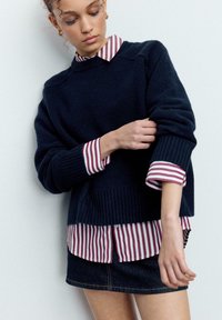 Mornarsko modra pletena majica z rebrastimi manšetami, nošena preko bordo in bele črtaste srajce s srajčnimi gumbi, kombinirana s temno denim krilo.