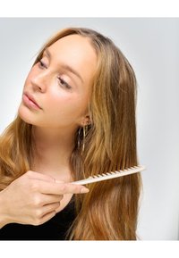 Grzebień plastikowy z szerokimi zębami, w jasnobeżowym kolorze, trzymany w dłoni, przesuwany przez długie, proste, jasnobrązowe włosy. Widoczna gładka faktura.
