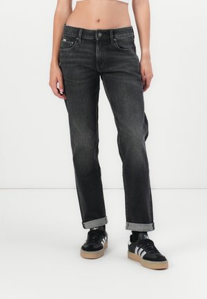 Person trägt schwarze, gerade geschnittene Jeans mit Umschlag und schwarze Sneakers mit weißen Streifen, steht vor einem weißen Hintergrund.