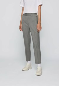 Pantalons rayés gris avec une taille ajustée et deux poches avant zippées. Le tissu a une texture lisse. Associés à des baskets blanches et des chaussettes.