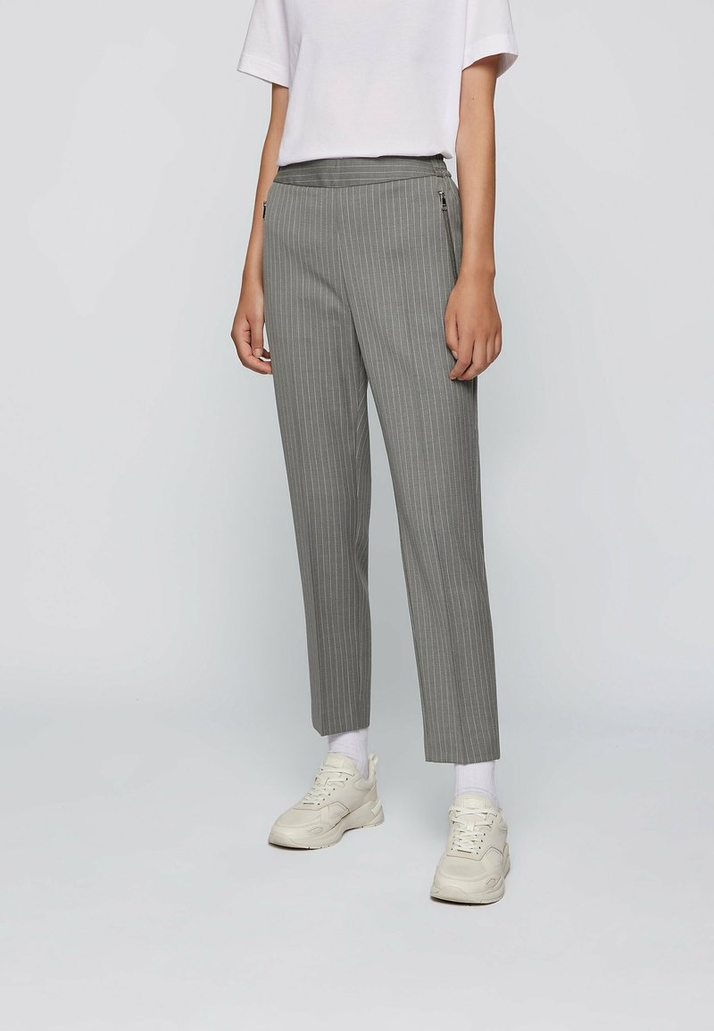 Pantalons rayés gris avec une taille ajustée et deux poches avant zippées. Le tissu a une texture lisse. Associés à des baskets blanches et des chaussettes.