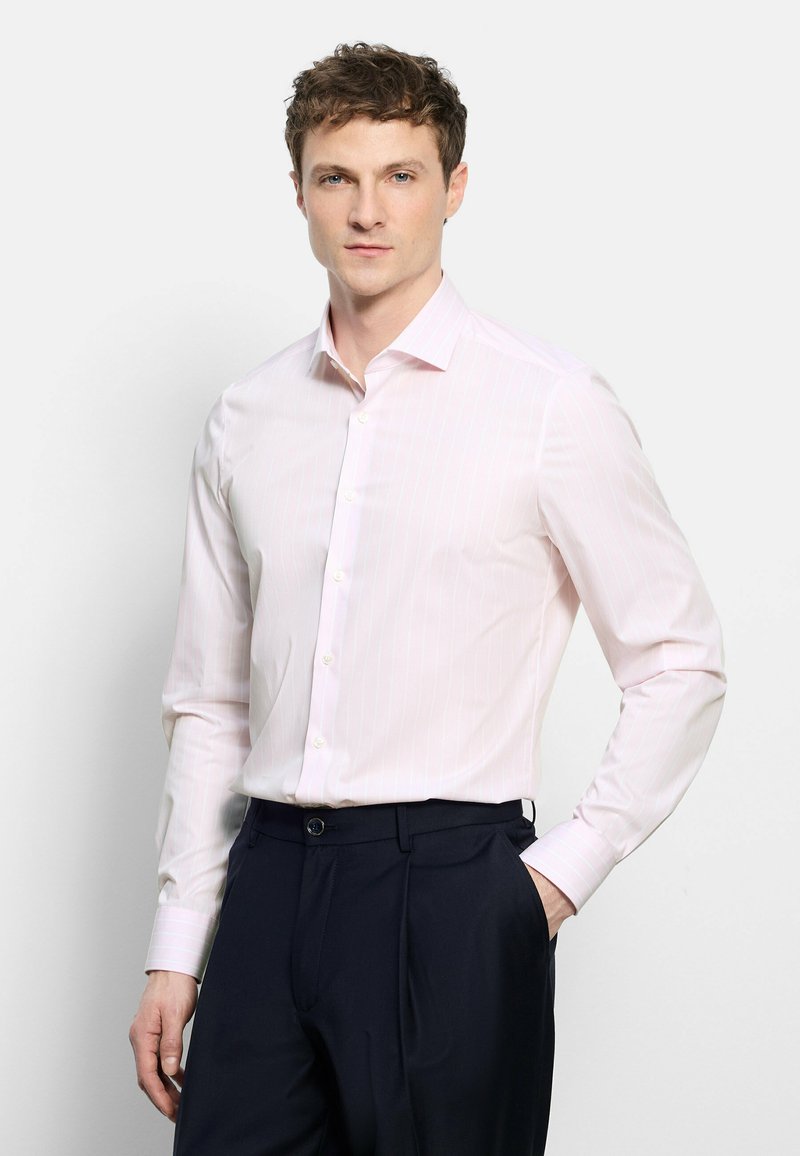 Jeune homme portant une chemise rose clair boutonnée rentrée dans un pantalon bleu marine foncé, debout avec une main dans la poche sur fond blanc.