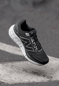 Chaussure de sport noire avec une tige en maille texturée, des accents réfléchissants et une semelle blanche rembourrée avec un motif en vagues. Les lacets sont noirs.