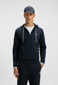 Felpa con zip di colore blu navy con lacci bianchi e logo. Il materiale appare liscio. Abbinata a pantaloni coordinati e un berretto scuro con logo.