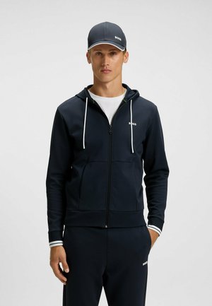 Marinblå zip-up hoodie med vita dragsnören och en logotyp. Materialet verkar slätt. Matchad med byxor och en mörk keps med logotyp.
