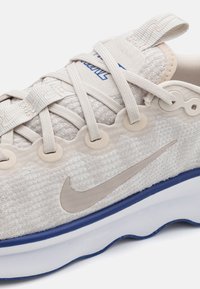 Lys beige sportssko med en struktureret stofoverdel, beige accenter, blå detaljer og en sål med bølgemønster. Har flade snørebånd og logo.