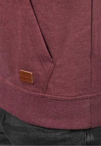 Närbild av en vinröd hoodie-ficka med synlig sömnad och en liten brun läderlogga ovanför den ribbade midjan.