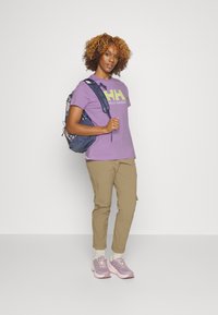 Camiseta morada con logo de HH, pantalones caqui y zapatillas rosas. La mochila tiene una base azul oscura con un diseño estampado.