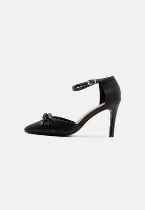 Chaussure noire à talon haut pointu avec bride à la cheville et petit nœud sur le bout, sur fond blanc.