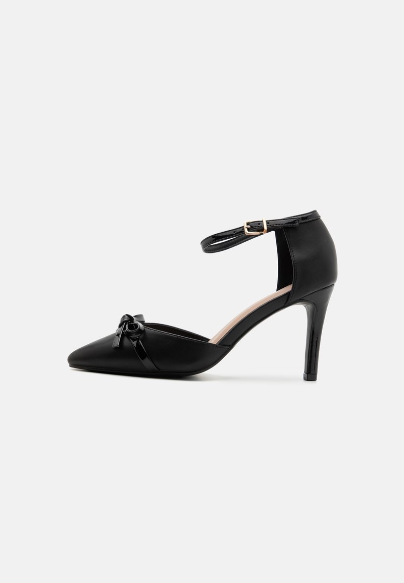 Chaussure noire à talon haut pointu avec bride à la cheville et petit nœud sur le bout, sur fond blanc.