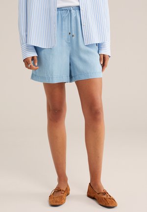 Žena oblečená v svetlomodrých šortkách so sťahovacou šnúrkou, pruhovanom modro-bielom tričku a hnedých mokasínach, stojaca pred neutrálnym pozadím.