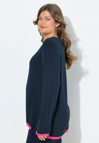 Femme aux longs cheveux ondulés portant un pull bleu marine avec des finitions roses, se tenant de profil et souriant devant un fond blanc.