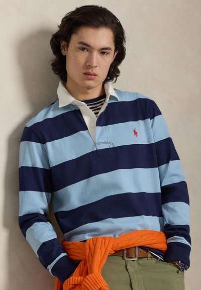 Polo a maniche lunghe a righe blu e blu navy con colletto bianco, con un piccolo logo rosso. Maglione arancione annodato in vita.