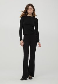 Maglietta nera a maniche lunghe e pantaloni svasati, entrambi in maglia. Tessuto liscio con silhouette aderente. Il modello indossa calzature nere. Sfondo neutro.