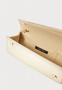 Kultainen metallilompakko, jossa on sileä pinta, napitus suljenta, sisätasku ja musta etiketti, jossa lukee "ANNA FIELD."