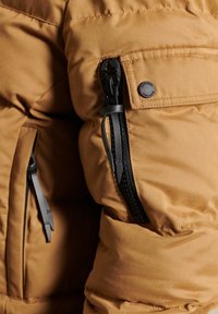 Veste matelassée marron à texture mate, dotée d'une poche zippée, d'une fermeture éclair noire et d'un bouton circulaire de marque sur l'épaule.