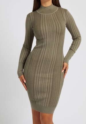 Femme portant une robe tricotée ajustée, côtelée, à manches longues, de couleur vert olive avec un col montant, debout devant un fond gris uni.
