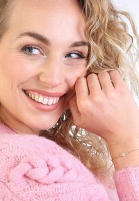 Gouden armband met een kleine heldere steen, gedragen aan een hand met lichte huid. De persoon heeft golvend blond haar en draagt een roze trui.