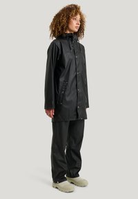 Mustaharjoitettu musta vesitiivis sadetakki, jossa on huppu, nepparipainikkeet ja sivutaskut, yhdistettynä samanvärisiin mustiin housuihin ja vaaleanvihreisiin jalkineisiin.