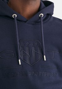Granatowa bluza z kapturem wykonana z bawełny, z embosowanym logo i tekstem, z sznurkami i metalowymi końcówkami. Projekt zawiera wzór tarczy.