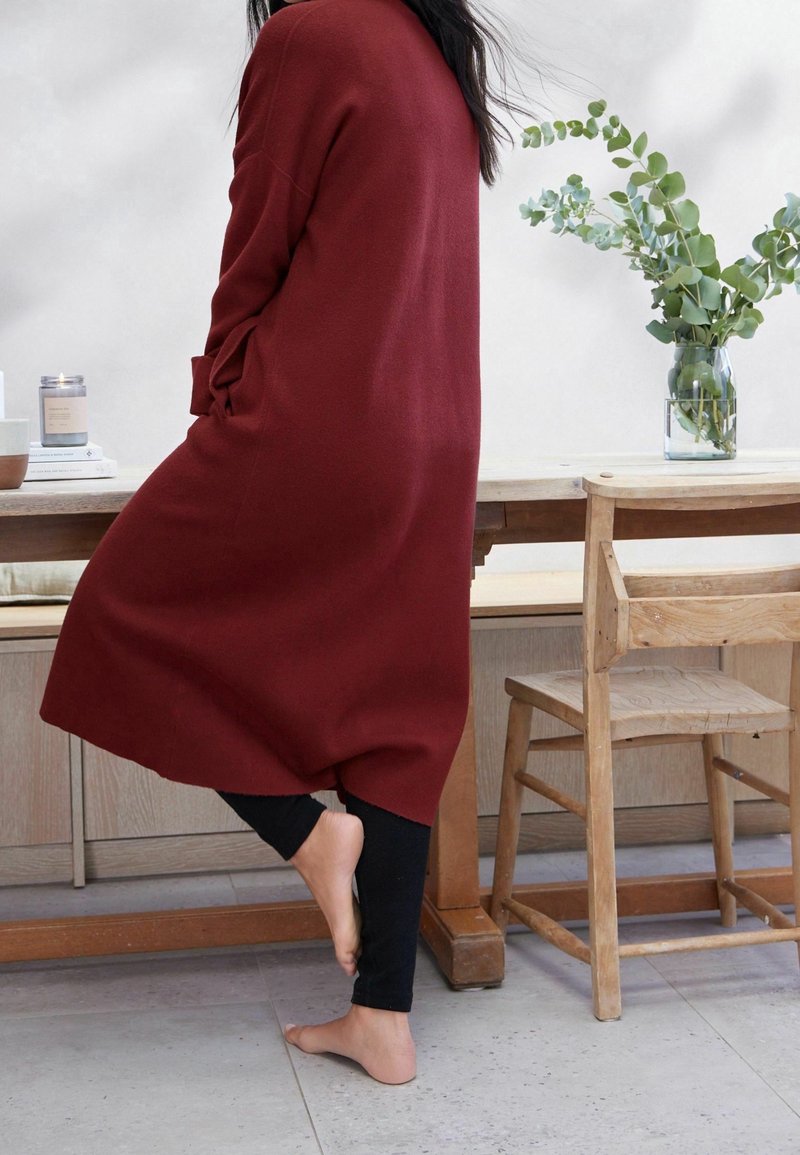 Personne debout pieds nus sur un pied à côté d'une table et d'une chaise en bois, portant un long cardigan rouge et des leggings noirs à l'intérieur.
