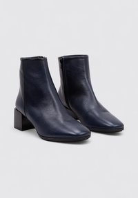 Botas de cuero azul marino hasta el tobillo con punta afilada, tacón en bloque y detalle de cremallera lateral. Textura suave con un diseño minimalista.