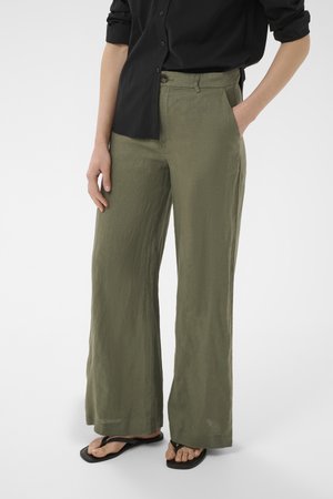 Personne portant un pantalon large vert olive, une chemise noire boutonnée et des tongs noires, avec une main dans la poche.