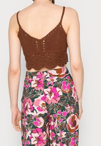 Hnědý háčkovaný crop top s vlněným lemem a nastavitelnými ramínky, spárovaný s kalhotami s květinovým vzorem v růžových, zelených a hnědých tónech.