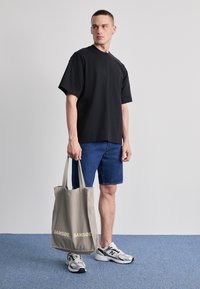 Tricou negru cu manșon scurt, pantaloni scurți din denim, geantă de umăr gri cu inscripția "SAMSØE" și adidași gri/albi pe o podea texturată albastră.