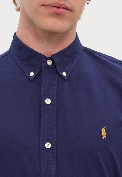 Camisa azul marinho com botões, com colarinho pontiagudo, apresentando botões brancos e um logótipo bordado a ouro no lado esquerdo do peito. Tecido de algodão suave.