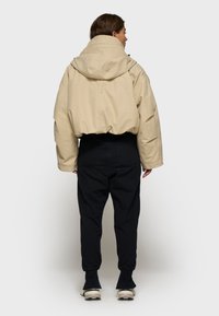 Kvinde vender væk iført en beige cropped hættetrøje, sorte joggingbukser og hvide sneakers mod en ensfarvet lys baggrund.