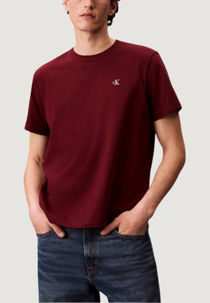 Uomo che indossa una semplice t-shirt a maniche corte bordeaux con un piccolo logo "cK" bianco sul petto, con le mani in tasca, abbinata a jeans blu.