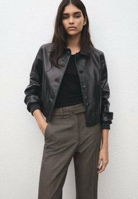 Młoda kobieta o długich ciemnych włosach, ubrana w czarną skórzaną zapinaną na guziki kurtkę, czarny top i brązowe spodnie z wysokim stanem oraz paskiem.