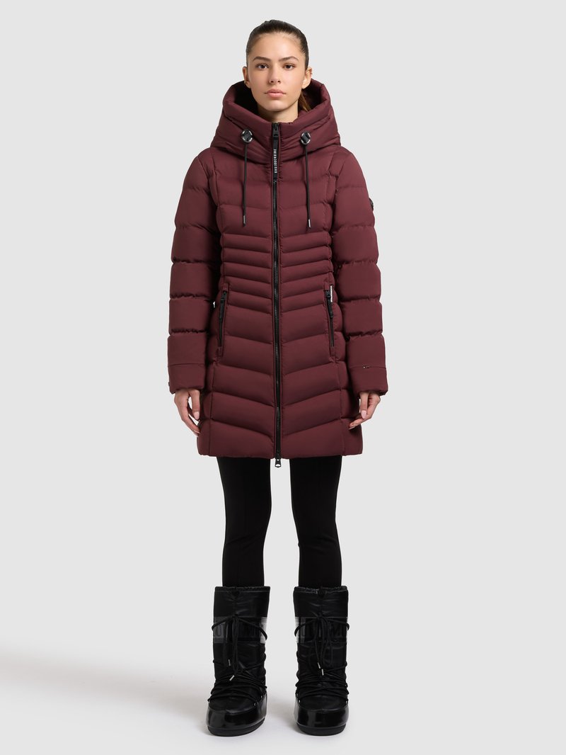 Giacca a piumino color marrone scuro con cappuccio alto, chiusura con zip e cuciture a chevron. Abbinata a stivali da neve neri con lacci elastici e drappeggio.