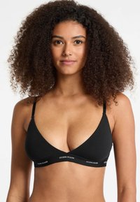 Black triangle bralette made of soft fabric with a wide band featuring "GUESS JEANS" text. Adjustable shoulder straps, smooth texture.