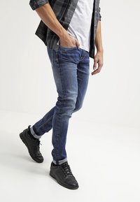 Jeans ajustados de mezclilla azul con un lavado desteñido, combinados con zapatillas negras tejidas y una camiseta gris bajo una camisa de cuadros, mostrados en una pose de caminata.