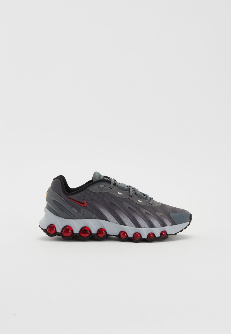 Zapato deportivo gris con acentos rojos y parte superior texturizada. Cuenta con una suela acolchada con bolsas de aire rojas visibles y un diseño elegante y moderno.