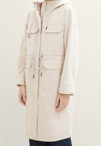 Abrigo largo beige con capucha, dos bolsillos frontales, cintura ajustable con cordón y cierre de cremallera. Textura suave y diseño minimalista.