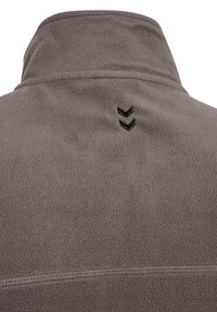 Nahaufnahme eines grauen Fleece-Stoffs mit einem hochgestellten Kragen und zwei kleinen schwarzen Chevron-förmigen Logos im oberen Rückenbereich.