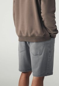 Pantaloni corti in cotone grigi con taglio dritto, dotati di due tasche posteriori e una superficie liscia. Completati da una felpa marrone.